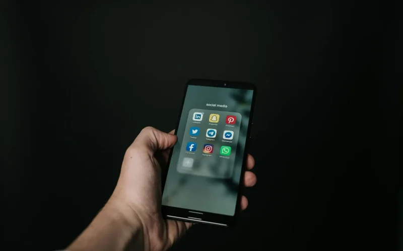 Close-up of a hand holding a smartphone displaying various social media app icons on a dark background.